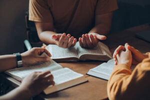 Christian family sitting around a wooden table with open bible page and holding each other's hand praying together